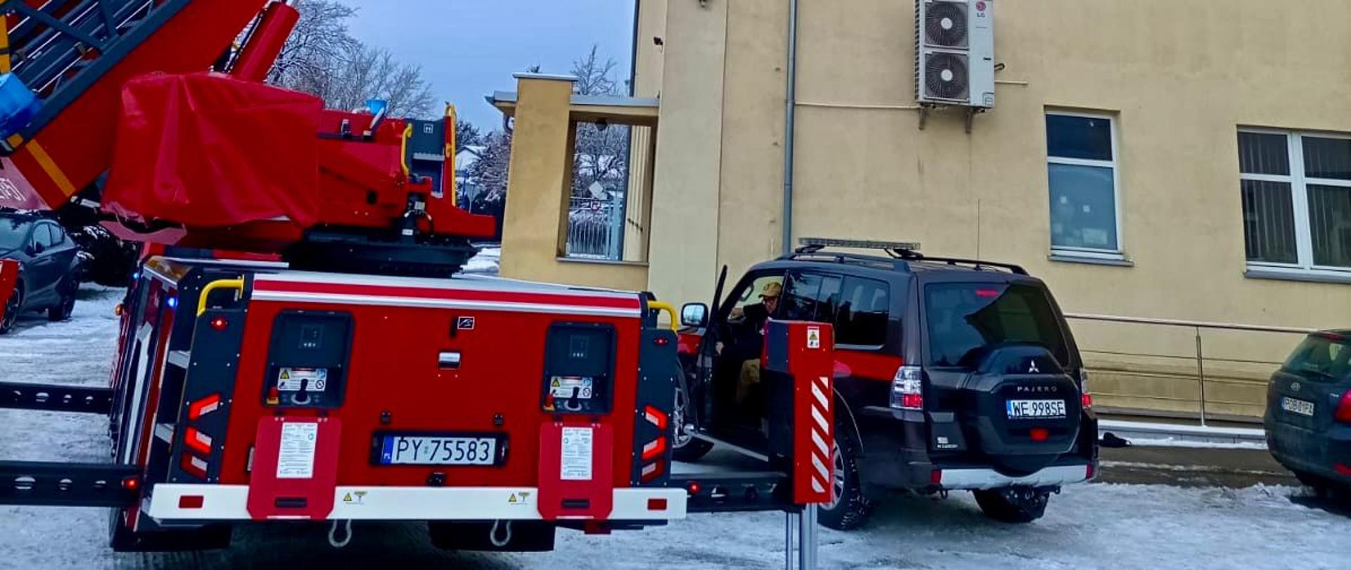 Czerwony wóz strażacki z wysuniętym długim podnośnikiem koszowym stoi na zaśnieżonym podwórzu przy budynku. Podnośnik sięga w górę wzdłuż ściany budynku. W tle widać inne budynki, drzewa oraz kilka osób i zaparkowane radiowozy.