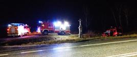 Zdjęcie przedstawia rozbity samochód osobowy w kolorze czerwonym który stoi poza jezdnią na trawie, ma otwarte tylnie lewe drzwi. Jest pora wieczorna. Po lewej stronie na drodze dojazdowej do drogi głównej DK 94 stoją dwa czerwone samochody pożarnicze z włączonymi światłami błyskowymi. Jeden z nich ma włączone światła na zabudowie pożarniczej i oświetla teren zdarzenia. Po prawej stronie widać betonowa figurkę z krzyżem. Asfalt na drodze głównej jest mokry od padającego deszczu a po prawej stronie widać na asfalcie położony pachołek ostrzegawczy w kolorze pomarańczowym z białymi odblaskowymi paskami.