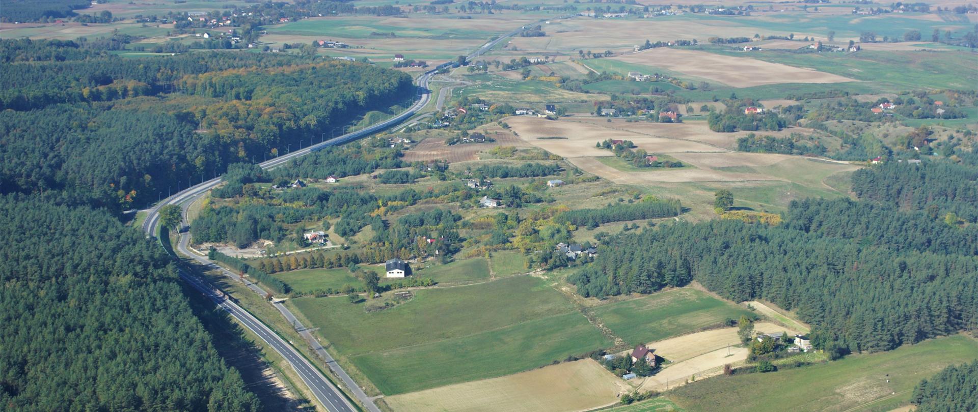 Zdjęcie ilustracyjne drogi krajowej przechodzącej przez tereny rolnicze i leśne. W centrum wstęga drogi, po lewej las, a po prawej krajobraz rolniczy z polami i gospodarstwami rolnymi.