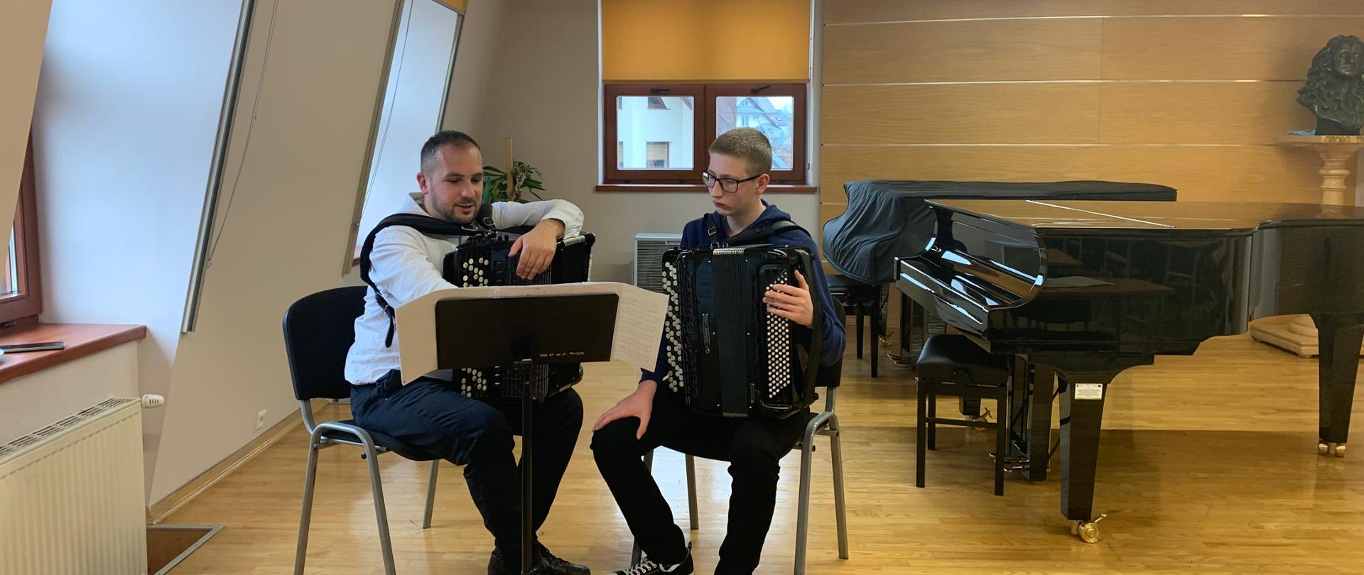 Warsztaty akordeonowe na sali kameralnej. Na pierwszym planie prowadzący warsztaty z uczniem, przed nimi pulpit z nutami, siedzą na krzesłach trzymając na kolanach akordeony. Po lewej stronie kaloryfer, dwa okna, za nimi okno a pod nim nawilżacz powietrza oraz palma doniczkowa. Po prawej stronie dwa fortepiany, jeden z nich przykryty pokrowcem
