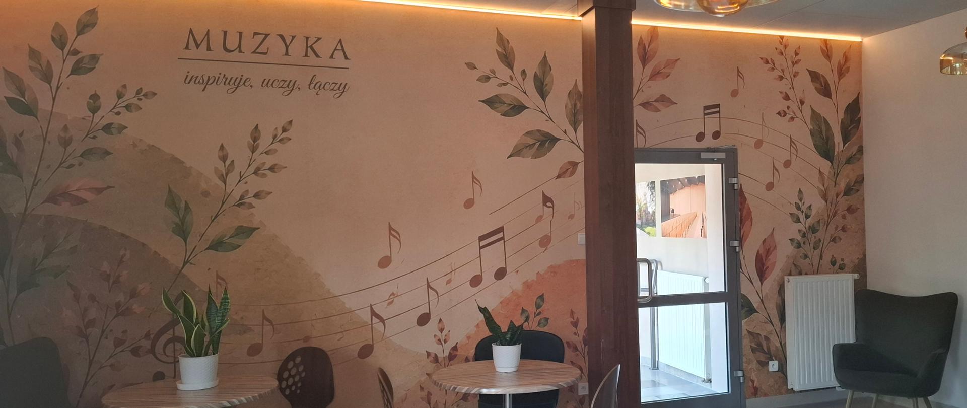Jasna przestrzeń z okrągłymi stolikami i kolorowymi krzesłami. Na ścianie znajduje się duża ilustracja roślin i instrumentów muzycznych oraz napis „MUZYKA – inspiruje, uczy, łączy”.