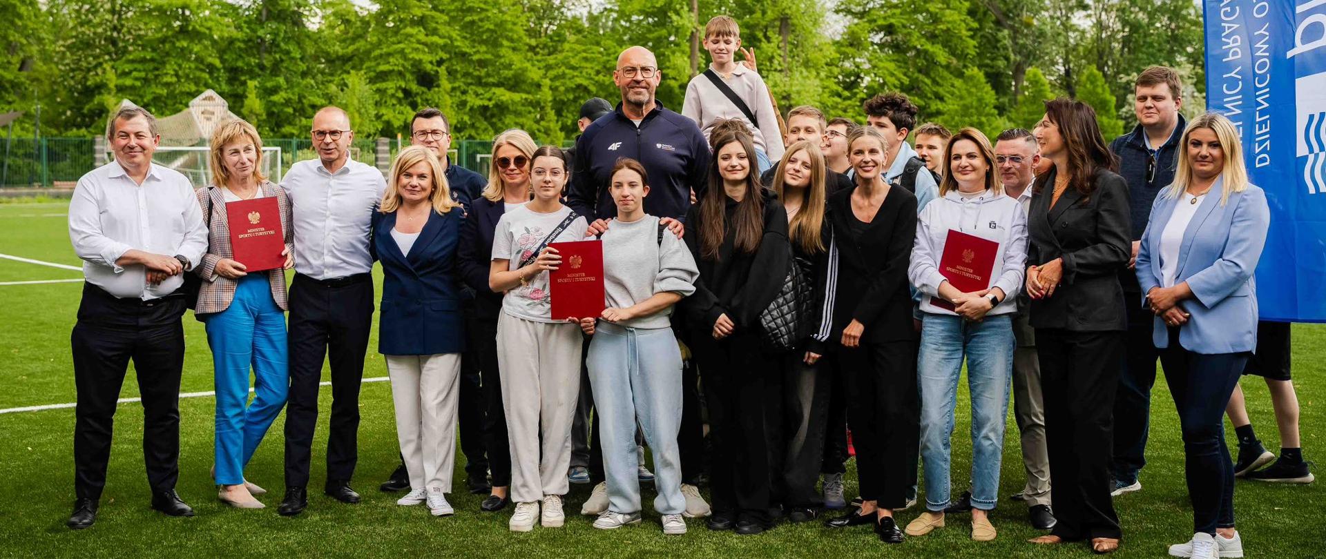 Zdjęcie zbiorowe na boisku piłkarskim. Minister sportu i turystyki Jakub Rutnicki, młodzież i dorośli pozują do zdjęcia z decyzją ministra o dofinansowaniu budowy pełnowymiarowego boiska piłkarskiego.