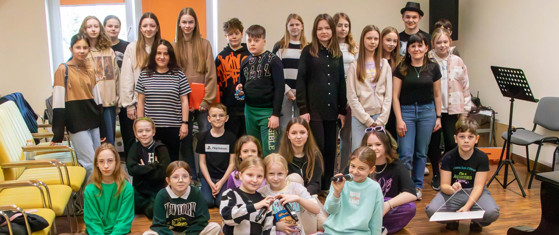 Na zdjęciu grupa kilkunastu dzieci, wśród nich dwie nauczycielki. Część osób stoi, część klęczy, część siedzi na podłodze w auli szkolnej. Po bokach widać puste krzesła.