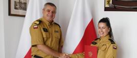 Strażak w mundurze musztardowym ściska dłoń strażaczce w mundurze musztardowym która trzyma czerwoną teczkę za nimi stoją dwie flagi Polski na ścianach wiszą obraz oraz szabla.