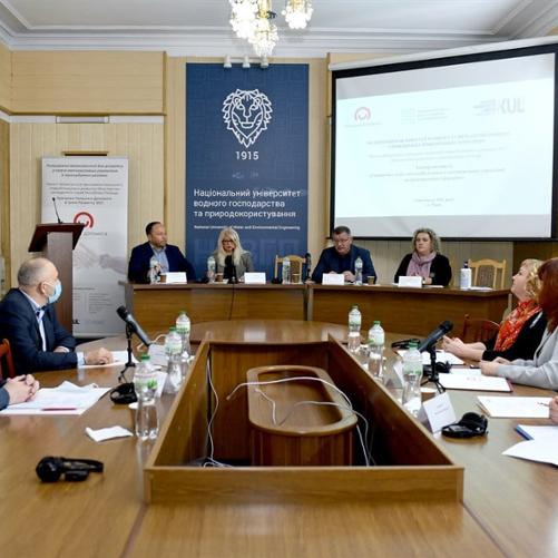 Grupa ludzi siedzi przy okrągłym stole i przy mównicy w eleganckim pomieszczeniu. 