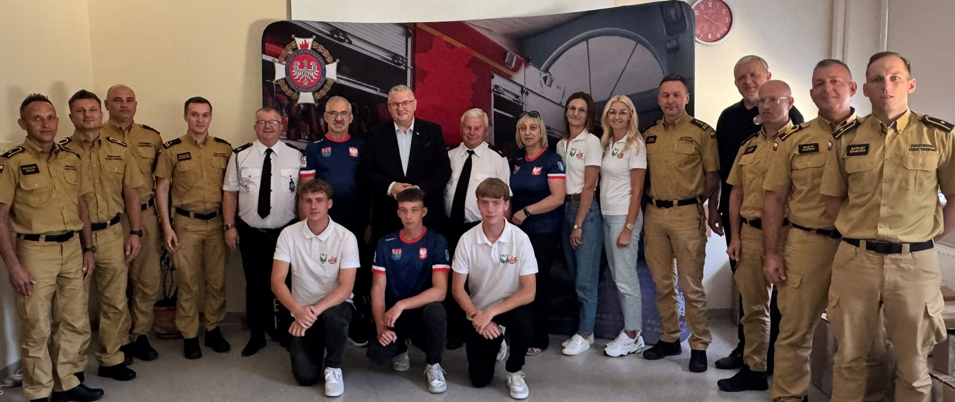 Grupa strażaków, organizatorów i młodzieży pozuje do wspólnego zdjęcia podczas wydarzenia. Większość uczestników ubrana jest w mundury lub koszulki z emblematami strażackimi. W tle znajduje się ścianka z grafiką samochodu pożarniczego i logo Związku Ochotniczych Straży Pożarnych.