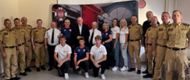 Grupa strażaków, organizatorów i młodzieży pozuje do wspólnego zdjęcia podczas wydarzenia. Większość uczestników ubrana jest w mundury lub koszulki z emblematami strażackimi. W tle znajduje się ścianka z grafiką samochodu pożarniczego i logo Związku Ochotniczych Straży Pożarnych.