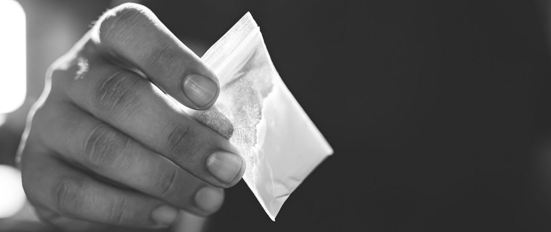 Czarno-białe zdjęcie. Zbliżenie na dłoń trzymającą woreczek strunowy z białym proszkiem.