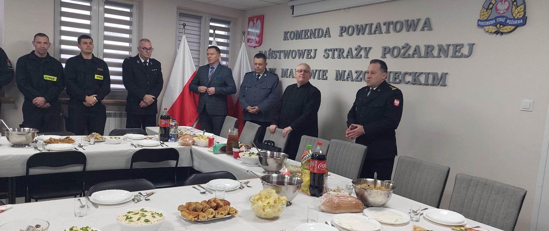 Komendant przemawia podczas spotkania wigilijnego. Obok stołów stoją funkcjonariusze oraz zaproszeni goście