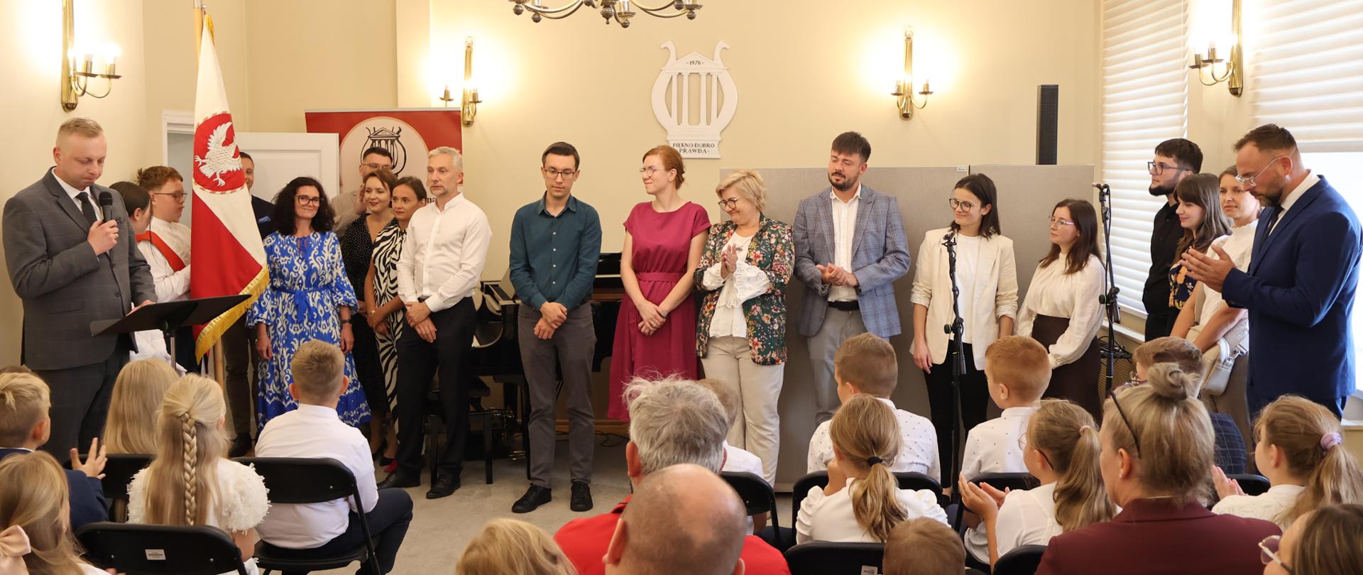 Grupa nauczycieli stojących przed uczniami i rodzicami siedzącymi na krzesłach w auli szkoły.