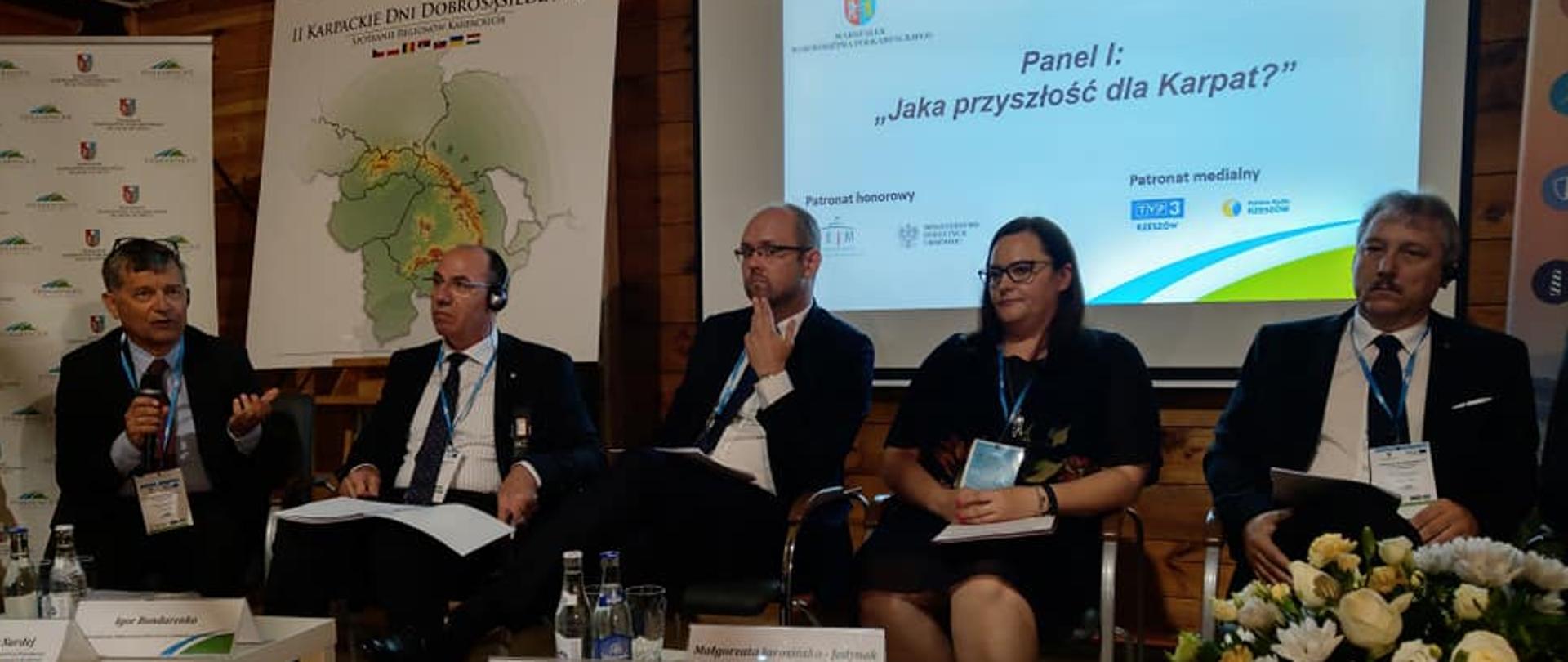 Pięć osób siedzi na krzesłach, za nimi baner z nazwą wydarzenia oraz ekran z tytułem panelu dyskusyjnego.