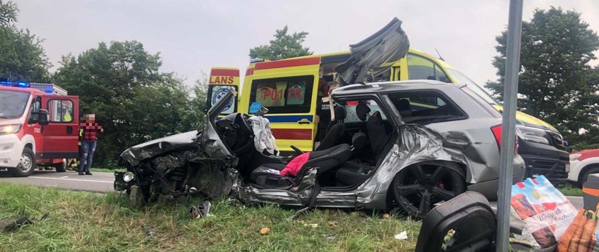 Dzień, wrak rozbitego auta po działach straży pożarnej, w tle ambulans.
