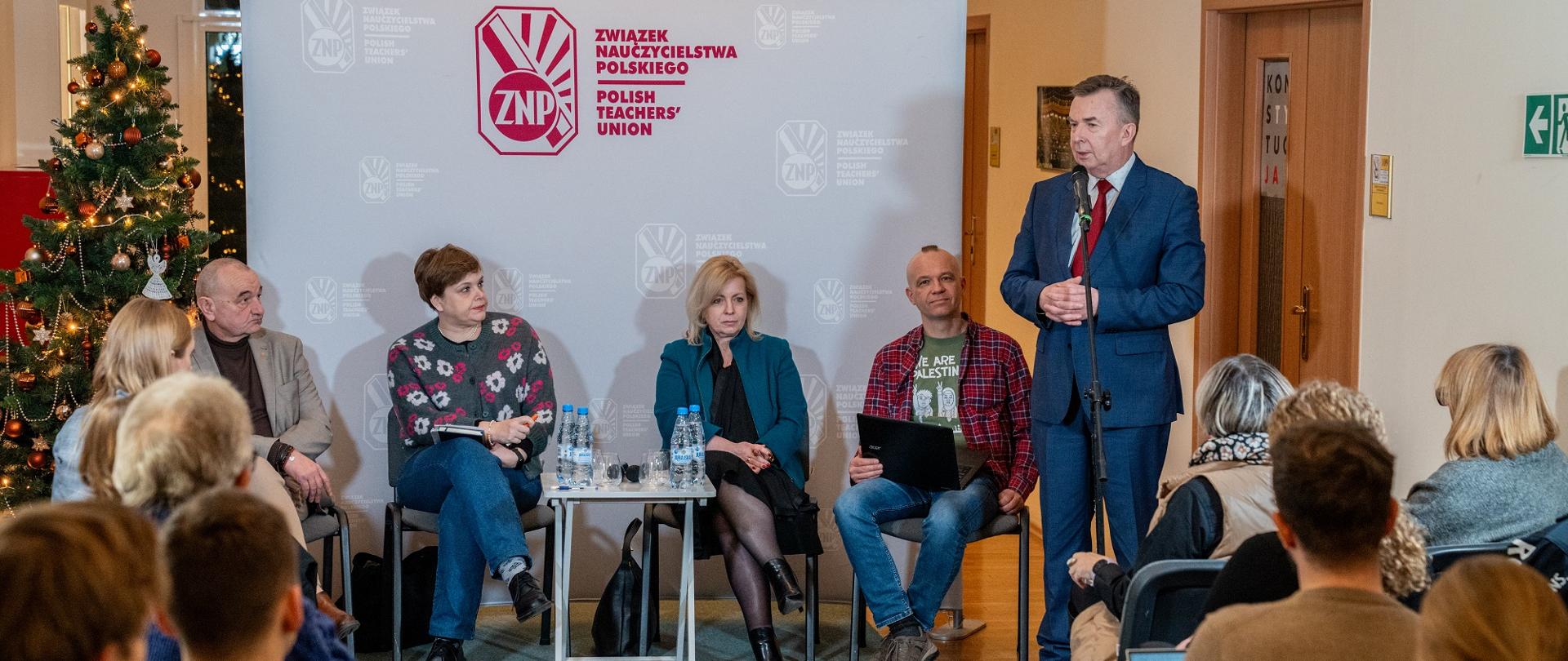 W sali na ustawionych w rzędy krzesłach siedzą ludzie, przed nimi pod ścianką z napisem Związek Nauczycielstwa Polskiego siedzą cztery osoby, z boku stoi minister Wieczorek i mówi do mikrofonu na stojaku.