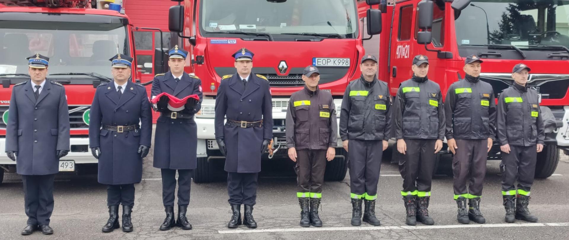 Uroczysta zbiórka przed budynkiem Komendy Powiatowej PSP w Opocznie. Umundurowani strażacy oraz poczet flagowy na tle samochodów pożarniczych.