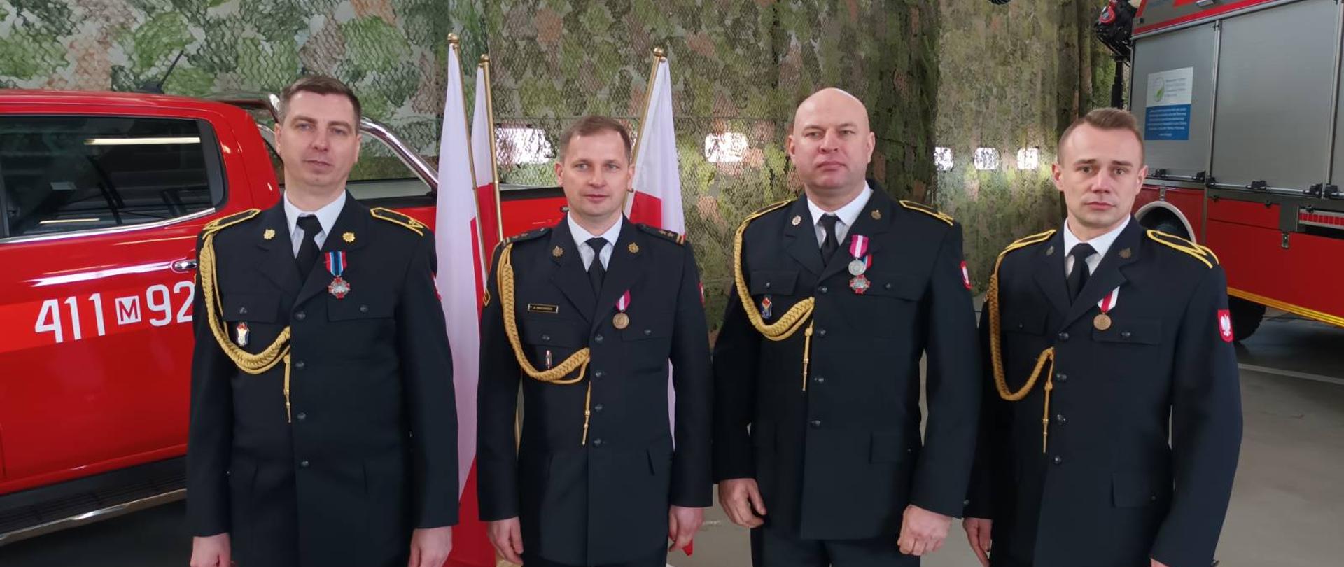 Uroczysty apel w KP PSP w Ciechanowie z udziałem Pana Macieja Wąsika wiceministra SWiA oraz gen. brygadiera Andrzeja Bartkowiaka komendanta głównego PSP - na zdjęciu 4 strażaków w ubraniu galowym po uroczystości. Stoją w garażu JRG za nimi stoją czerwone samochody strażackie.