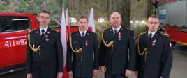 Uroczysty apel w KP PSP w Ciechanowie z udziałem Pana Macieja Wąsika wiceministra SWiA oraz gen. brygadiera Andrzeja Bartkowiaka komendanta głównego PSP - na zdjęciu 4 strażaków w ubraniu galowym po uroczystości. Stoją w garażu JRG za nimi stoją czerwone samochody strażackie.