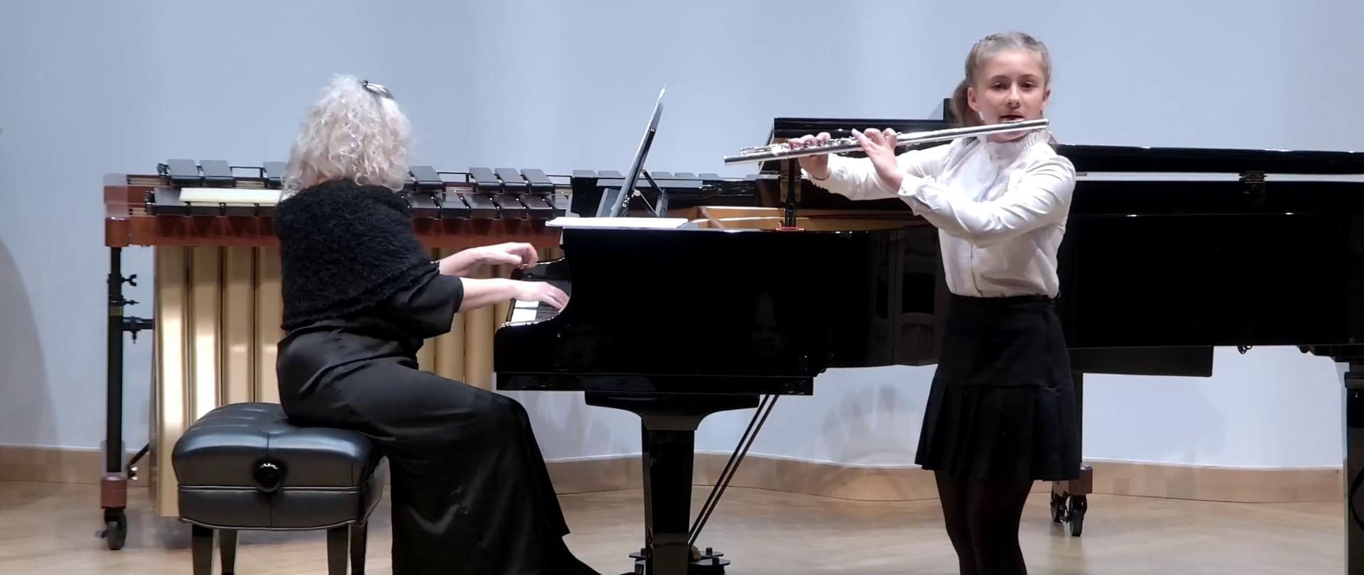 Zdjęcie. Na estradzie sali koncertowej występuje młoda flecistka z towarzyszeniem pianistki. Za fortepianem w tle stoi instrument perkusyjny - marimba.