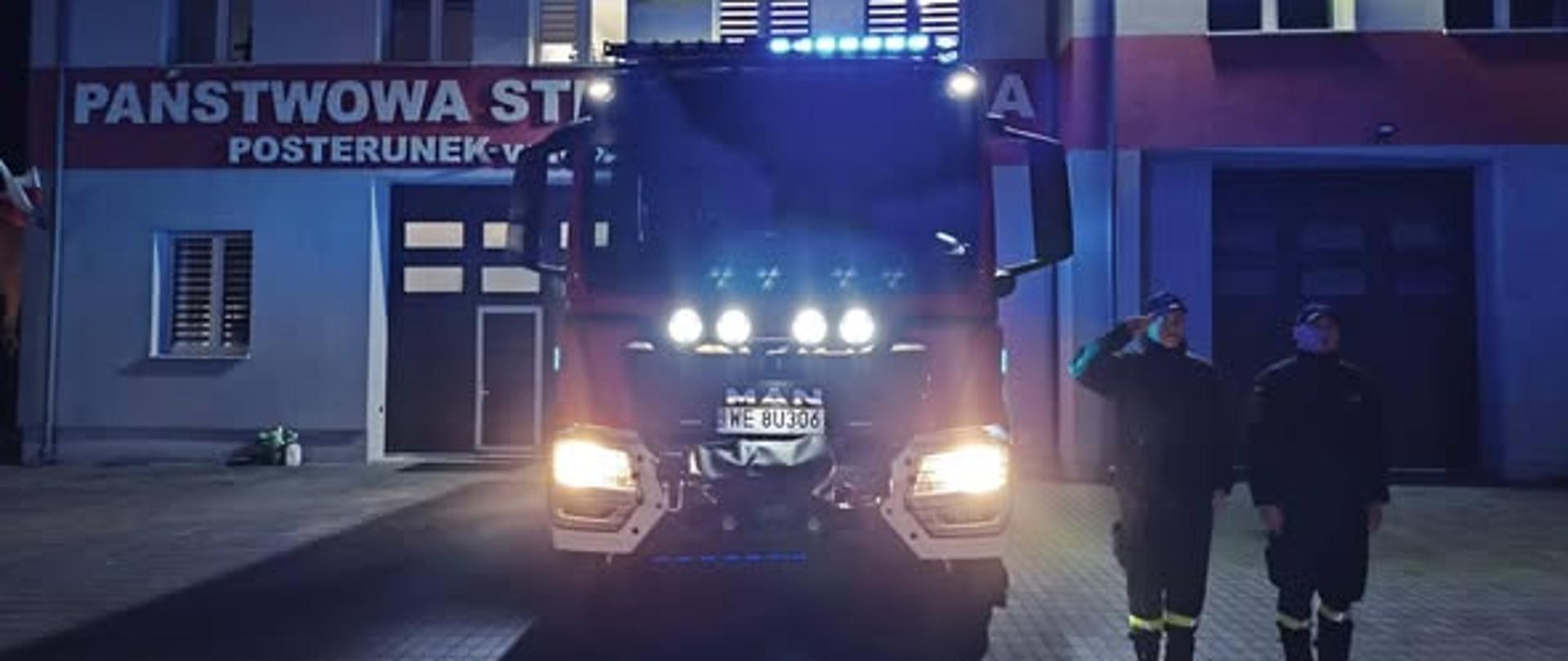 Dwóch strażaków stoi przy wozie strażackim i oddaje hołd zmarłemu druhowi z OSP w Jerzwałdzie.