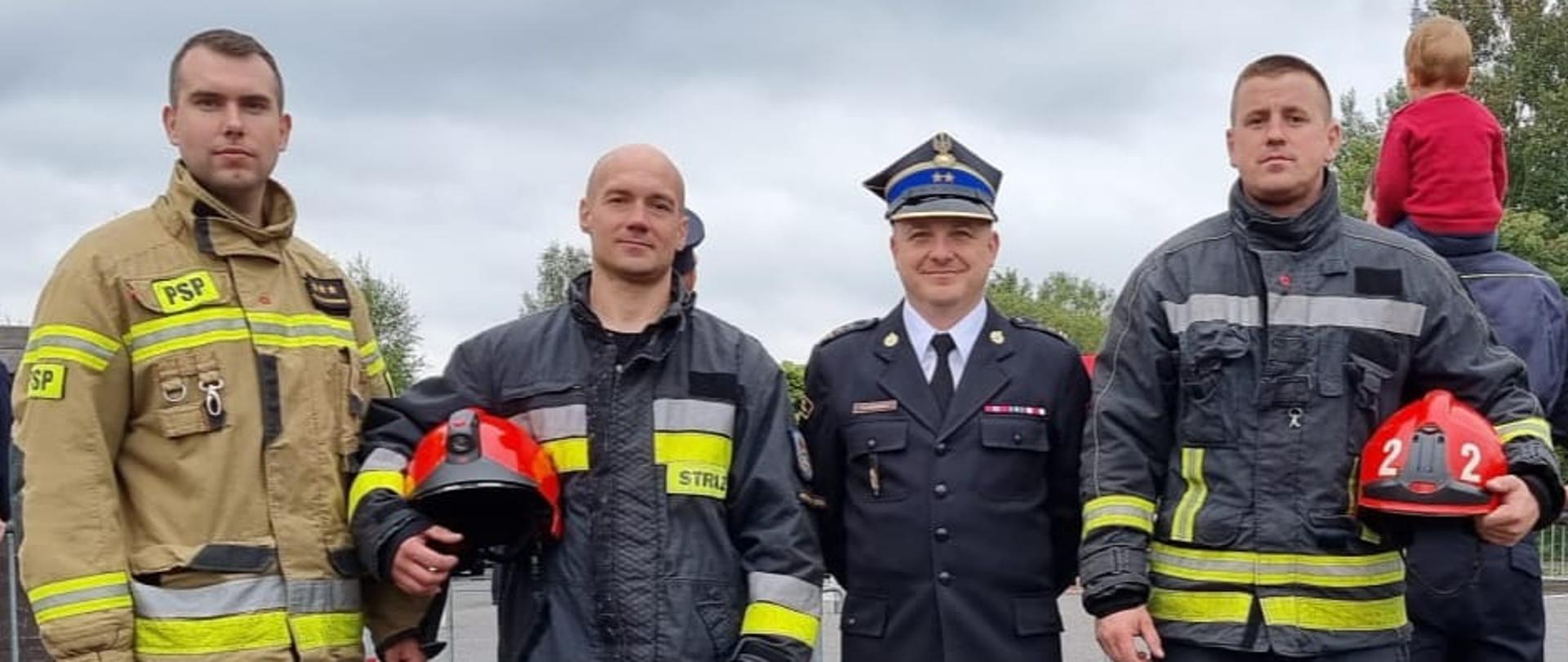 Na zdjęciu widać czterech strażaków, trzech z nich w umundurowaniu bojowym, jeden z nich w umundurowaniu galowym. Zdjęcie wykonane podczas zawodów TFA w Czechach w dzień. 