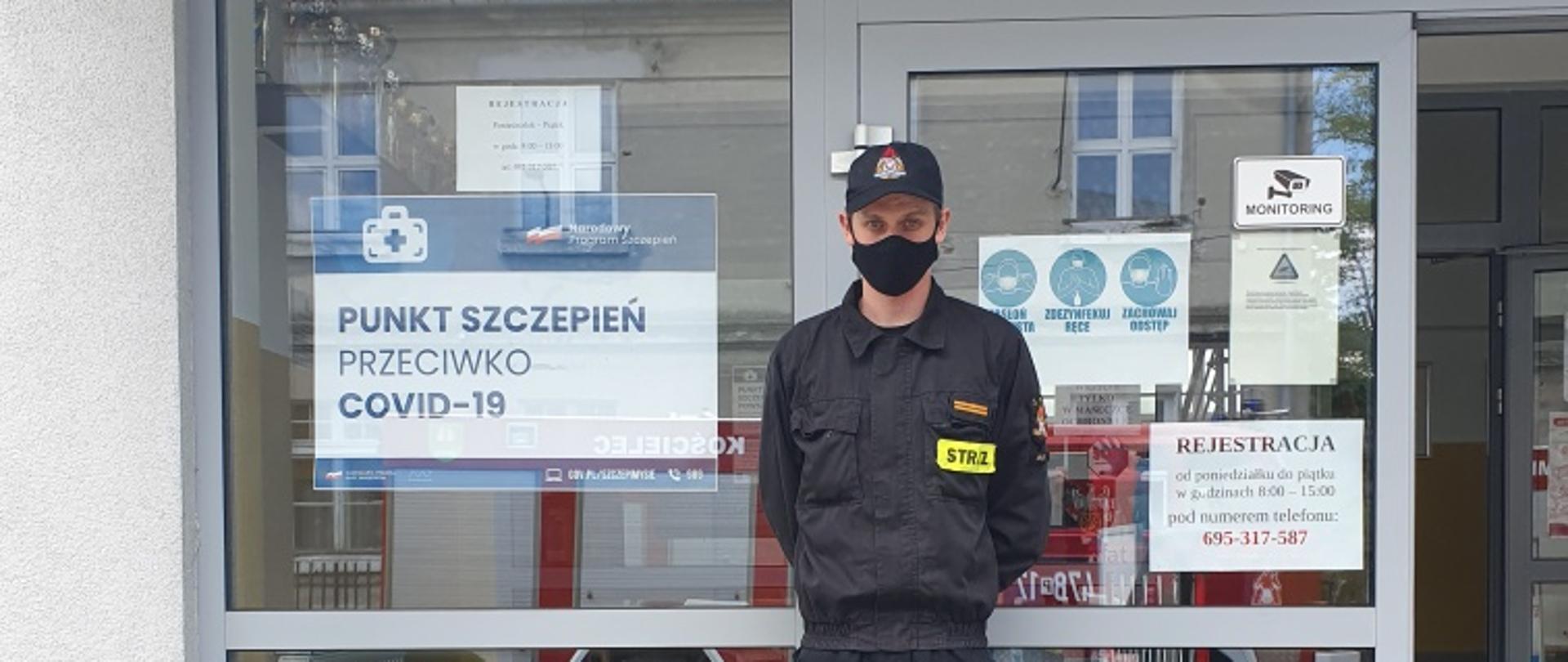 Zdjęcie przedstawia umundurowanego strażaka pomagającego przy punkcie szczepień. Strażak znajduje się przed budynkiem hali, w której zorganizowany jest punkt szczepień.