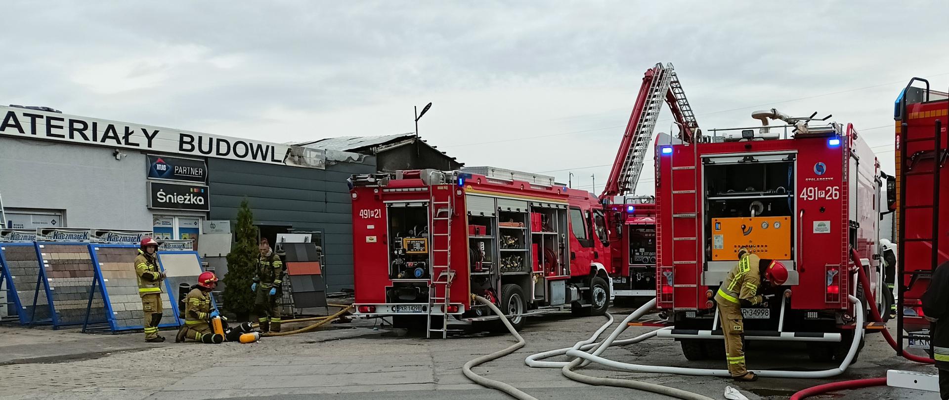 Zdjęcie przedstawia wozy strażacki biorące udział w zdarzeniu oraz strażaków odpoczywających po wyjściu ze strefy zagrożenia.