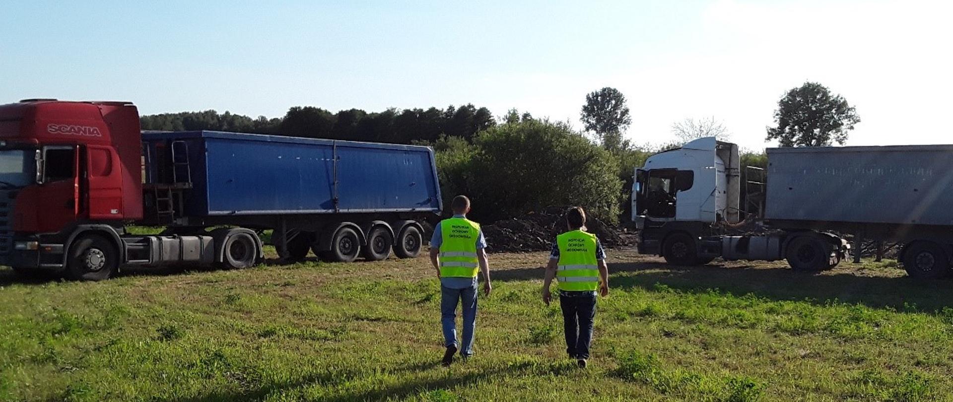 Dwóch inspektorów w zielonych kamizelkach z napisem „Inspekcja Ochrony Środowiska” w tle dwa ciągniki siodłowe z naczepami.