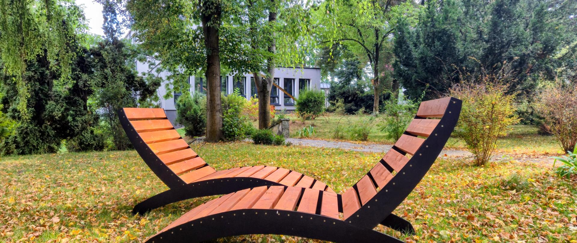 W ogrodzie na pierwszym planie stoją naprzeciw siebie dwa leżaki - z czarnym stelażem i drewnianymi szczebelkami. Na trawie leżą pożółknięte liście, w tle drzewa i krzewy oraz w oddali fragment szarego budynku.