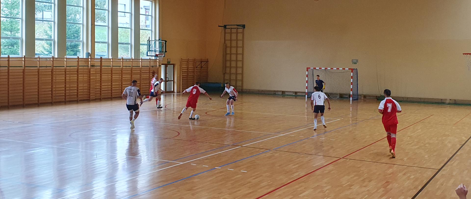 Na zdjęciu strażacy w strojach sportowych grający w piłkę nożna na parkiecie hali sportowej.