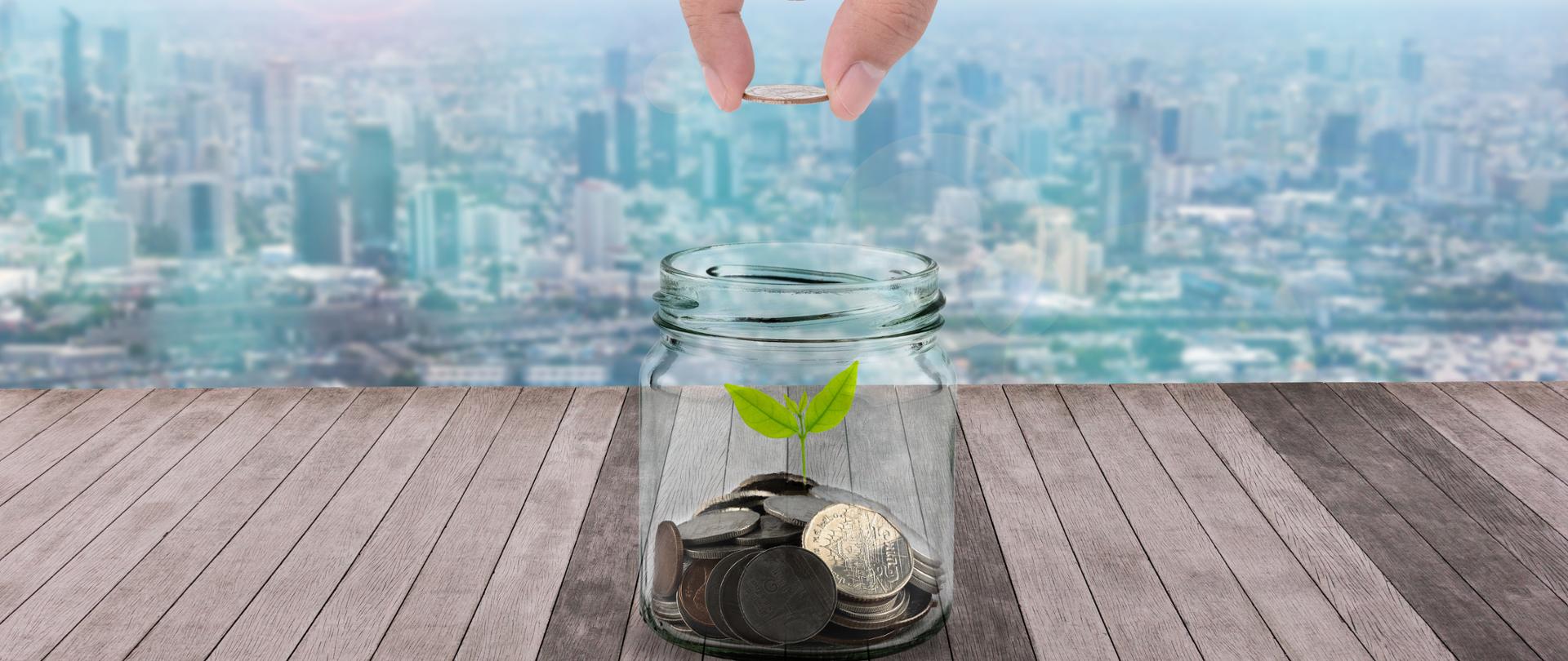 Dwa palce trzymające monetę nad słoiczkiem w którym widać monety i wychodzącą z nich listki rośliny na tle miasta 