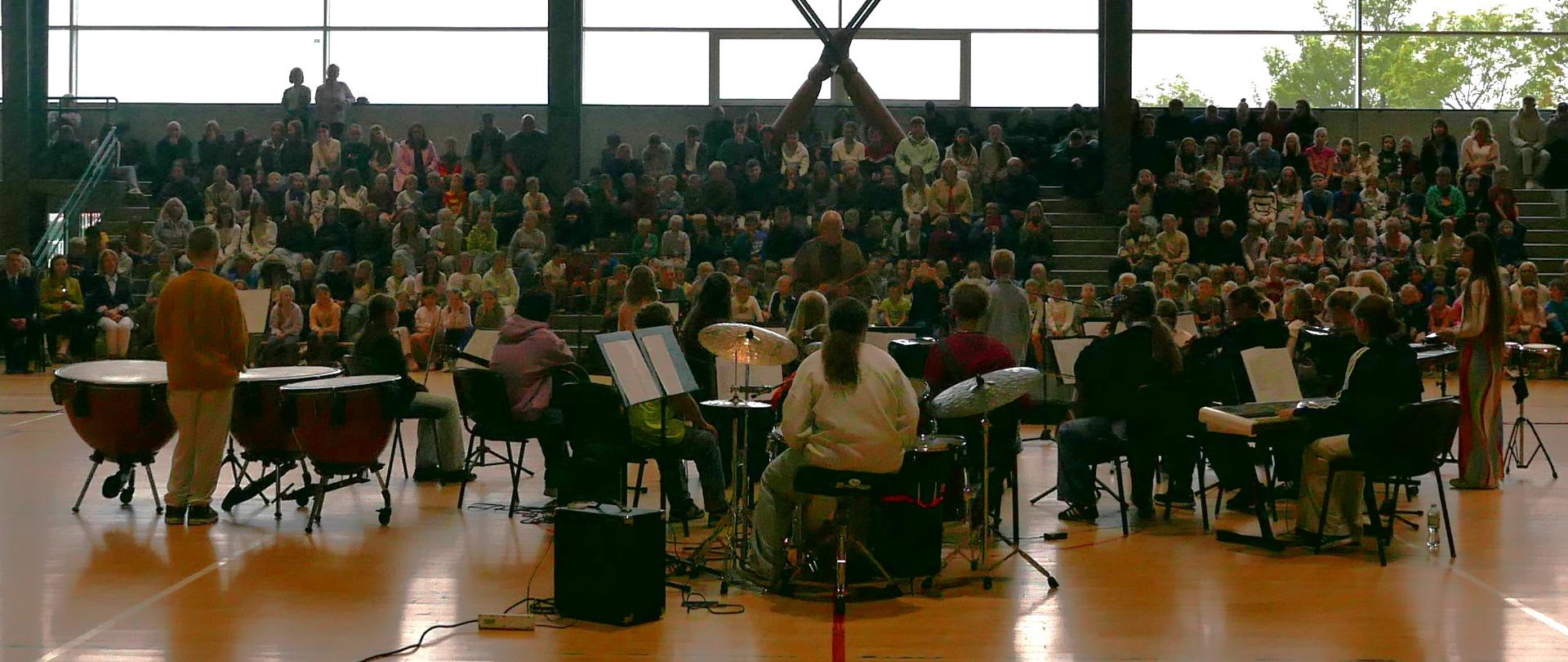 Inne ujęcie zespołu muzycznego. Na pierwszym planie orkiestra grająca koncert. Wszyscy siedzą twarzą do widowni za wyjątkiem dyrygenta. Dalej duża widownia złożona z młodzieży szkolnej. Za nimi ogromne okna hali sportowej.