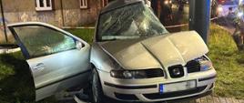 Zdjęcie przedstawia dramatyczne skutki wypadku samochodowego, rozgrywającego się w warunkach nocnych, na co wskazuje ograniczone oświetlenie. Widoczna mokra nawierzchnia chodnika przedstawia niedawne opady. Centralnym punktem kadru jest poważnie uszkodzony srebrny samochód osobowy, przypominający model Seat Toledo. Pojazd doznał rozległych zniszczeń, w tym wgniecionej maski i przedniego błotnika, całkowicie rozbitej i zapadniętej przedniej szyby oraz mocno zniekształconego dachu, który wydaje się być częściowo oderwany. Lewe drzwi są szeroko otwarte.
Samochód zatrzymał się tuż przy ciemnym, pionowym słupie, który wydaje się być nieuszkodzony lub jedynie minimalnie naruszony, co sugeruje, że to właśnie ten obiekt mógł być punktem kolizji. Wrak pojazdu stoi na chodniku. W tle, po prawej stronie, za słupem, widać fragment zieleni oraz niewyraźne sylwetki ludzi i prawdopodobnie innych pojazdów, w tym czerwony wóz strażacki, co wskazuje na obecność służb ratunkowych na miejscu zdarzenia.
Za miejscem wypadku rozciąga się miejski krajobraz, charakteryzujący się starszą zabudową. Widoczne są budynki mieszkalne z oknami, z których niektóre świecą, co dodaje scenie realizmu. Na ścianie jednego z budynków można dostrzec ślady graffiti. Cała scena jest słabo oświetlona, co potęguje wrażenie powagi sytuacji.
