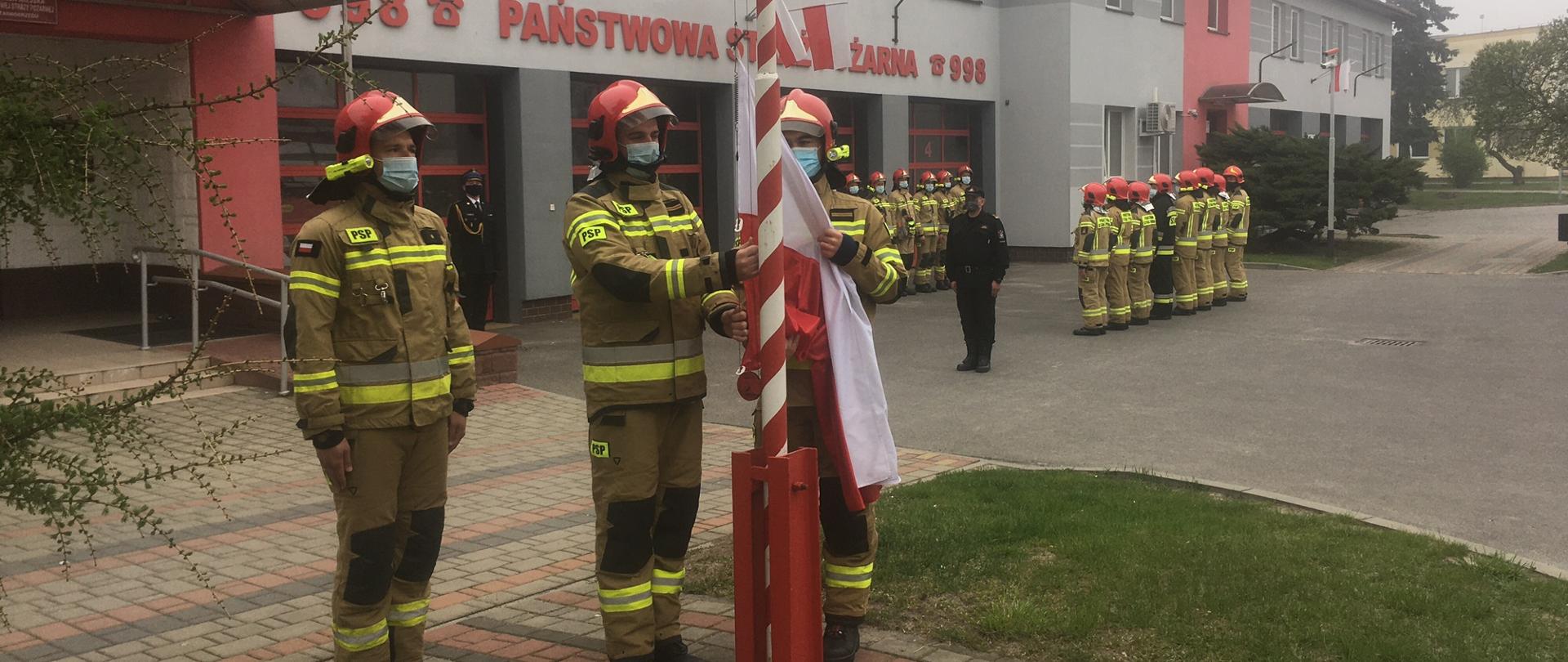 Trzech strażaków w umundurowaniu bojowym stoi przed masztem, jeden z nich zapina flagę. Na drugim planie, przed budynkiem stoją strażacy, którzy są ustawieni w dwóch szeregach.