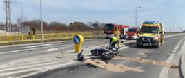 Zdjęcie przedstawia rozbity motocykl leżący na jezdni. Przy nim pochylony Policjant prowadzi czynności wyjaśniające, za nim znajduje się żółty ambulans oraz samochody Strazy Pożarnej 
