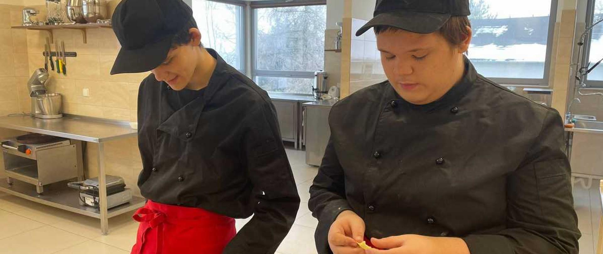 Zdjęcie przedstawia dwóch młodych kucharzy przy stole roboczym w profesjonalnej kuchni. Oboje ubrani są w czarne bluzy kucharskie i czerwone zapaski wiązane w talii. Na głowach mają czarne czapki typu bejsbolówka.
Osoba po lewej stronie jest pochylona nad blatem ze stali nierdzewnej, na którym leży rozwałkowane, jasne ciasto. Dłońmi formuje kawałki ciasta, prawdopodobnie lepiąc pierogi. Druga osoba, stojąca po prawej stronie, również trzyma w dłoniach kawałek ciasta i skupia się na jego formowaniu.
W tle widoczne jest wyposażenie kuchni, w tym metalowe regały ze sprzętami, okna z widokiem na zewnątrz oraz fragmenty innego blatu kuchennego i urządzeń gastronomicznych. Scena sugeruje szkolenie kulinarne lub warsztaty, podczas których uczestnicy uczą się przygotowywać tradycyjne dania.