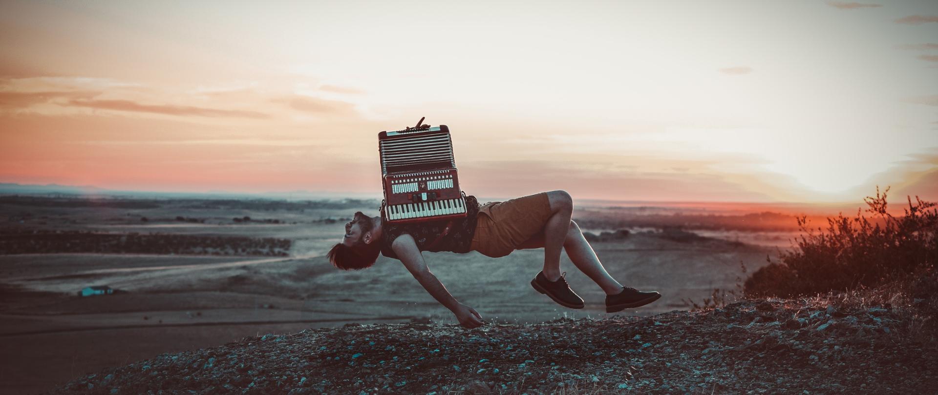 fotografia przedstawia ciemnowłosego mężczyznę z brodą, ubranego w ciemny podkoszulek i krótkie jasnobrązowe spodenki unoszącego się w powietrzu nad ziemią, trzymającego na torsie akordeon na tle zachodzącego słońca