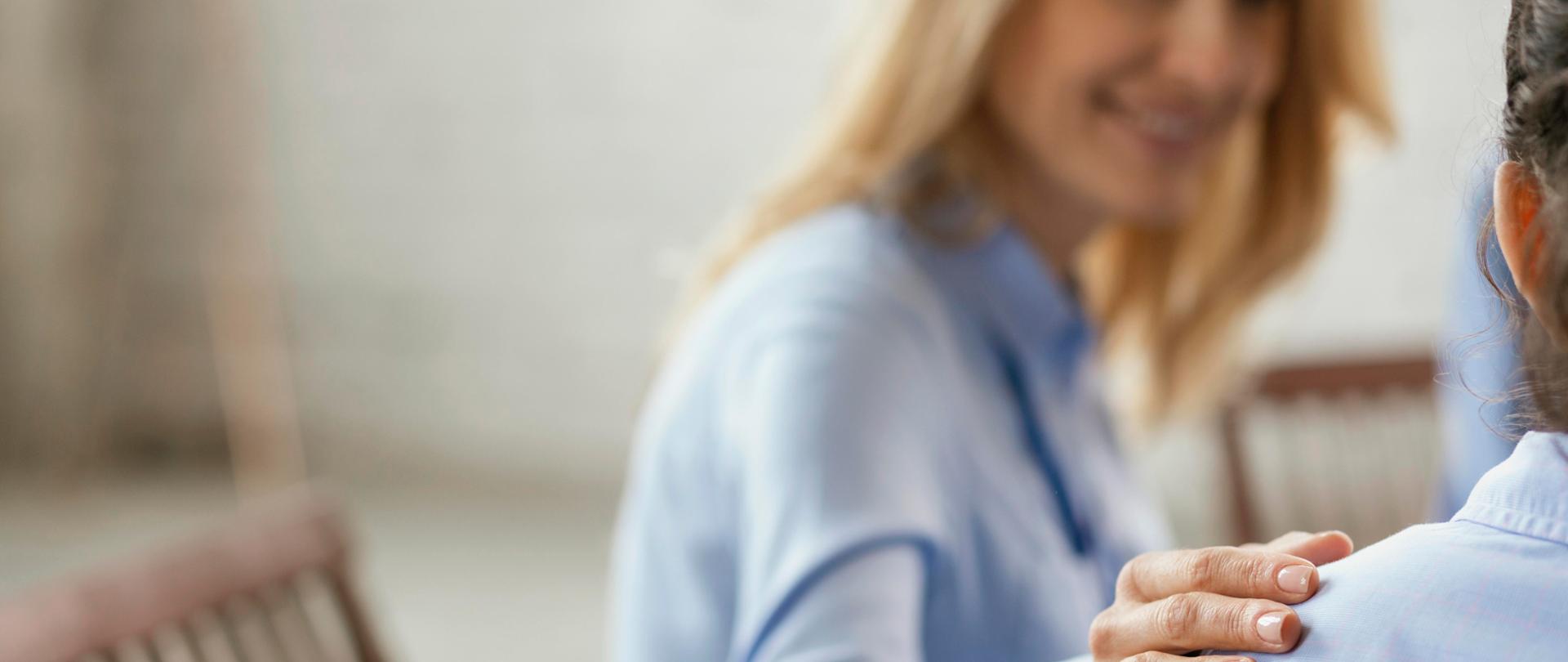 Częściowy wizerunek dwóch osób siedzących obok siebie na krzesłach. Jedna z osób trzyma dłoń na ramieniu drugiej. 