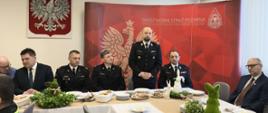 Przy stołach na których znajdują się dekoracje świąteczne i talerze, siedzą strażacy i zaproszenie goście. Strażacy ubrani są w mundury galowe zaproszeni goście odświętnie w garniturach. Jeden z zaproszonych gości w postawie stojącej przemawia. W tle logo Państwowej Straży Pożarnej oraz godło Polski i flagi