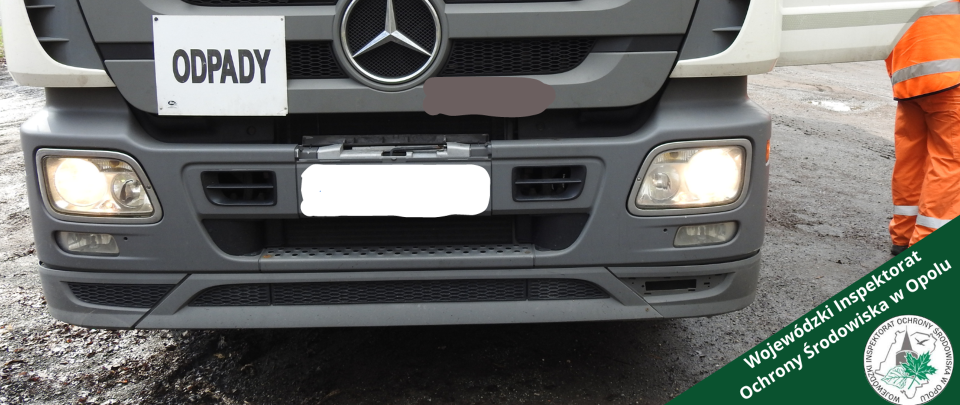 Przód samochodu ciężarowego przewożącego odpady. Dół samochodu w odcieniach szarości, góra biała. Obok znaku marki samochodu znajduje się tabliczka z napisem "odpady". Po prawej stronie zdjęcia widać otwarte drzwi od strony kierowcy. Obok otwartych drzwi widać nogi osoby ubranej w pomarańczowy strój składający się z długich spodni i kurtki z odblaskami.