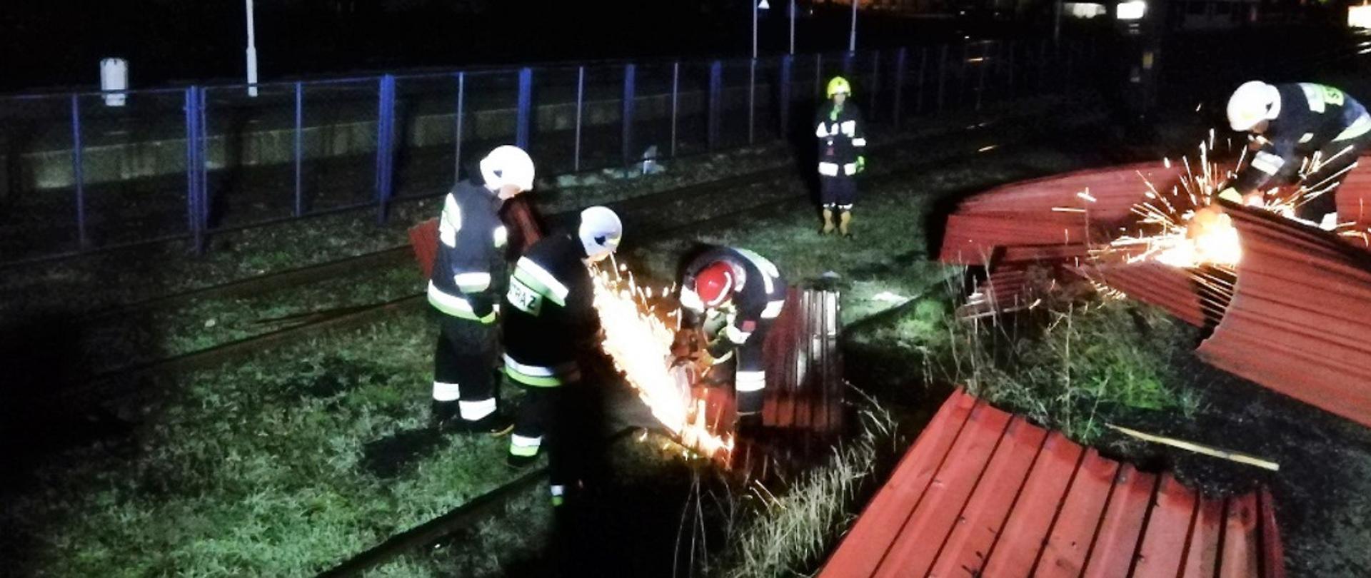 Na zdjęciu znajduje się trzech strażaków w pełnym uzbrojeniu, przy użyciu pilarek do cięcia stali usuwa zerwane arkusze blachy leżące na bocznicy kolejowe.