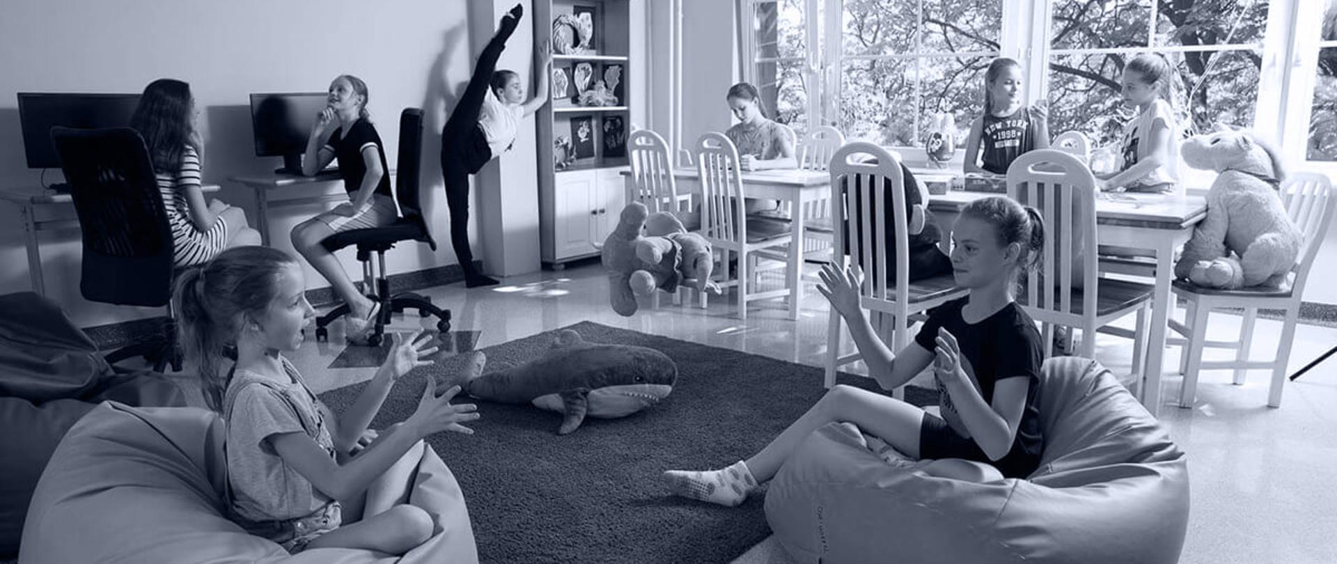 Na zdjęciu dzieci przebywające w świetlicy internatu - dwie dziewczynki siedzące przed komputerami, trzy odrabiające lekcje przy stole, jedna rozciągająca się oraz dwie siedzące na pufach i rozmawiające gestykulując 