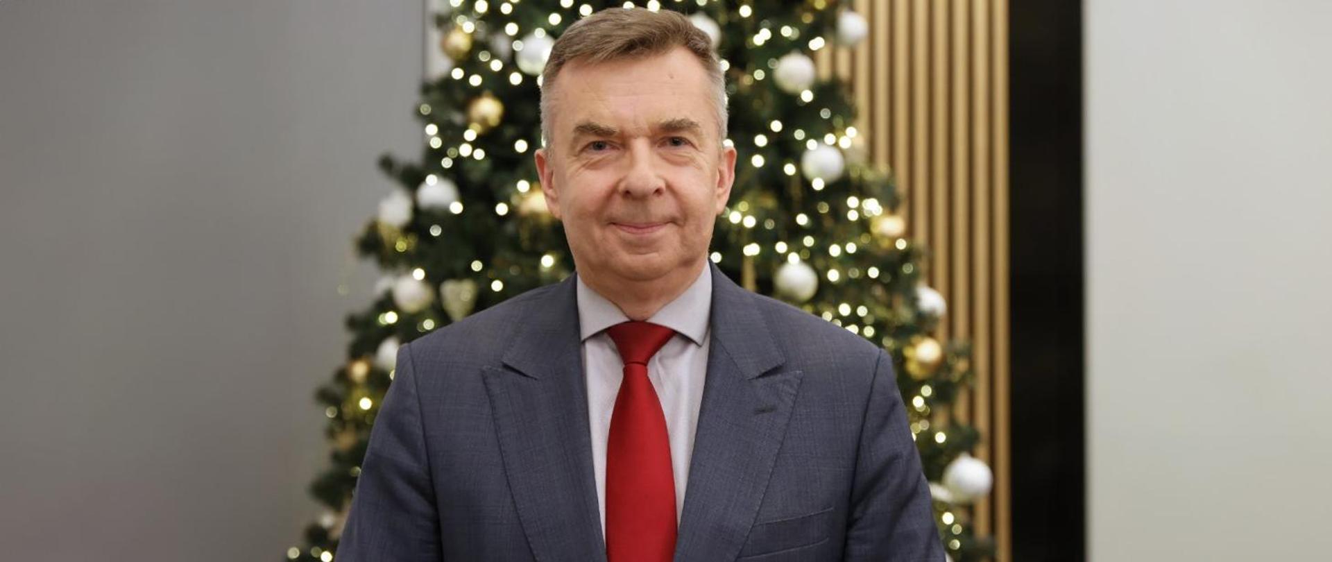 Minister Wieczorek stoi na tle udekorowanej bombkami i łańcuchami choinki.