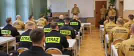 2. Przywitanie uczestników przez komendanta
Ujęcie sali wykładowej z perspektywy tylnej. Na pierwszym planie widoczne są plecy kursantów, ubranych w beżowe i czarne ubrania służbowe z napisami "STRAŻ" lub "PAŃSTWOWA STRAŻ POŻARNA". Z przodu sali, przed białym ekranem projekcyjnym, stoi oficer w beżowym mundurze strażackim, zwracający się w stronę grupy. Po prawej stronie w głębi stoi dwóch innych oficerów. Na ścianie wisi duży, okrągły zegar oraz tablice.
