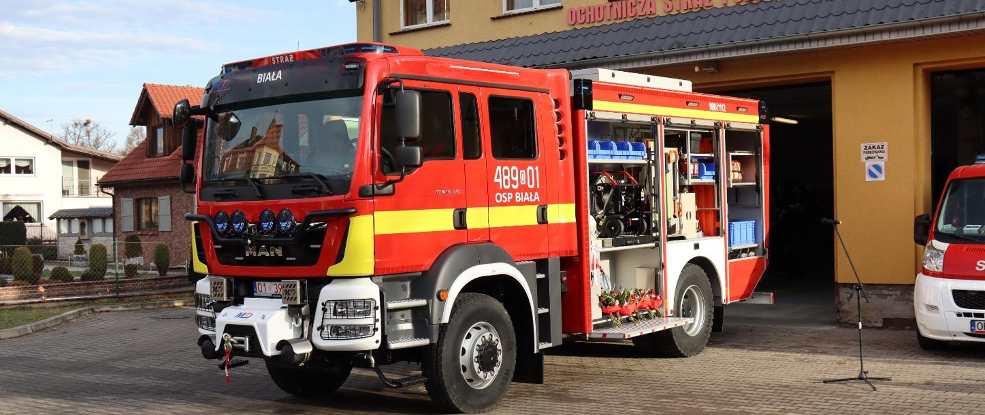 Na zdjęciu w centralnym miejscu widoczny samochód ratowniczo-gaśniczy marki MAN (kolor czerwony, białe zderzaki oraz nadkola oraz żółty pas przez całą długość kabiny). Pojazd ma otwarte skrytki i podesty obszaru roboczego. Na drzwiach przedziału załogi jest biały napis 489[o]01 a pod spodem OSP Biała. Pojazd stoi przodem do wyjazdu (widoczny przód i lewy bok) na kostce brukowej przed otwartą bramą remizy OSP Biała. Elewacja remizy pomalowana na kolor ciepły żółty z napisem "Ochotnicza Straż Pożarna w Białej" koloru czerwonego nad bramami wyjazdowymi. Po prawej stronie zdjęcia widoczny kawałek przodu innego samochodu strażackiego. Pomiędzy samochodami ustawiony na statywie mikrofon.
Po lewej stronie kadru widoczne zabudowania.