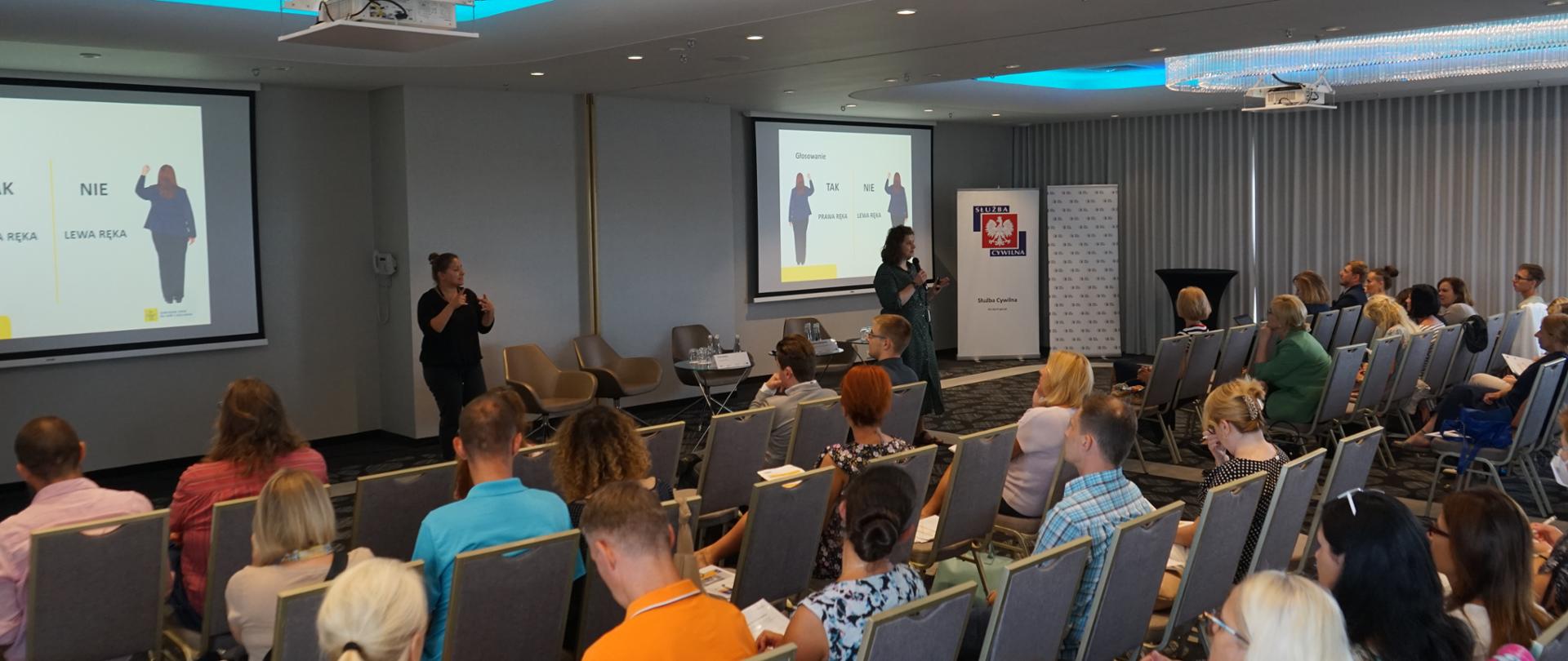Na sali konferencyjnej siedzi kilkadziesiąt osób, patrzą na dwie prelegentki, za nimi wyświetlana jest prezentacja.