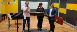 Na zdjęciu znajdują się pedagog prowadzący warsztaty - Wojciech Front oraz pani dyrektor PSM i nauczyciel kontrabasu w sali rytmicznej PSM w kolorach szaro, żółty, biały, na ścianie wisi godło i biała tablica w pięciolinię, po prawej stronie znajduje się szafa w kolorach szaro-żółtym, na ścianach wiszą kolorowe obrazki muzyczne, w tle na podłodze leży kontrabas