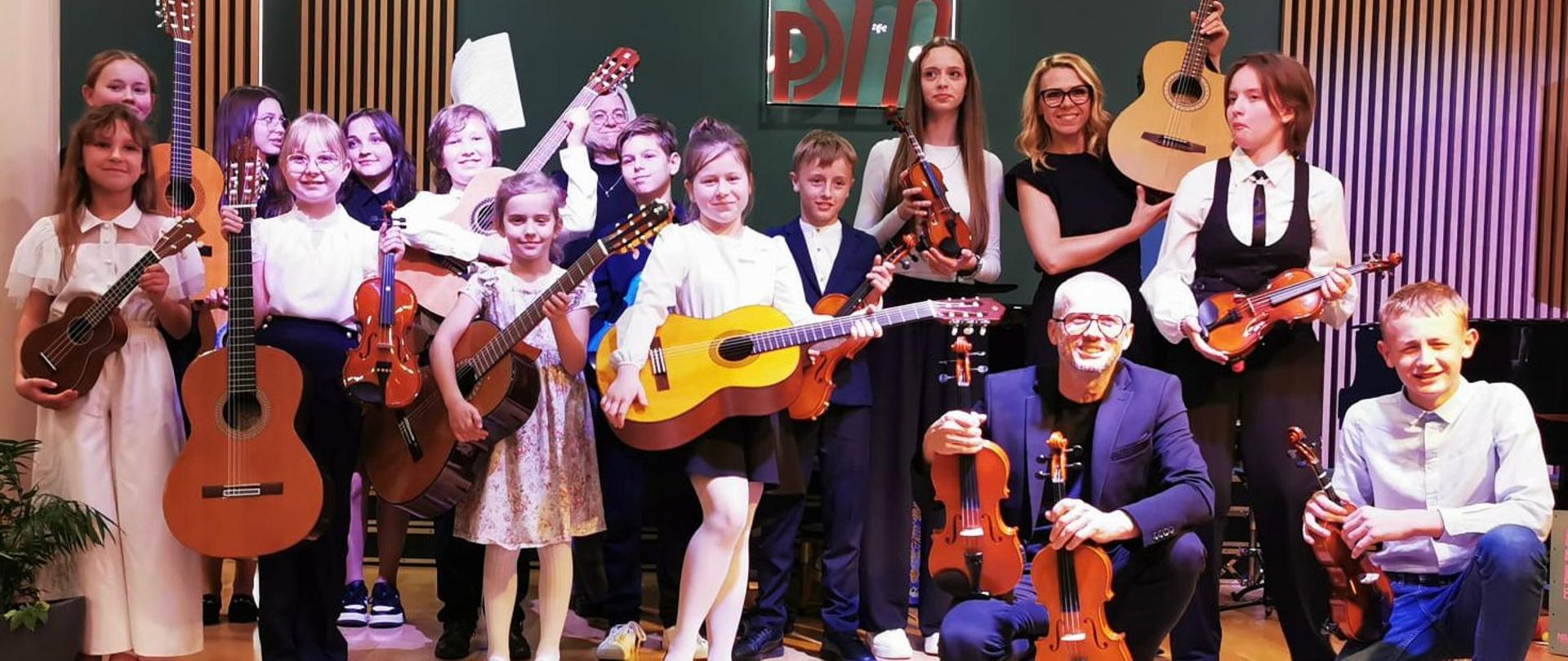 Na zdjęciu widać uśmiechnięte dzieci które w rękach trzymają skrzypce lub gitary niektórzy kucają. 