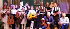 Na zdjęciu widać uśmiechnięte dzieci które w rękach trzymają skrzypce lub gitary niektórzy kucają. 