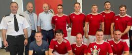 Drużyna w strojach piłkarskich pozuje do zdjęcia w dwuszeregu. Pierwszy rząd kuca, drugi stoi za nimi. Dwie osoby trzymają dyplom za zajęcie II miejsca i puchar. Razem z nimi stoi trzech organizatorów.