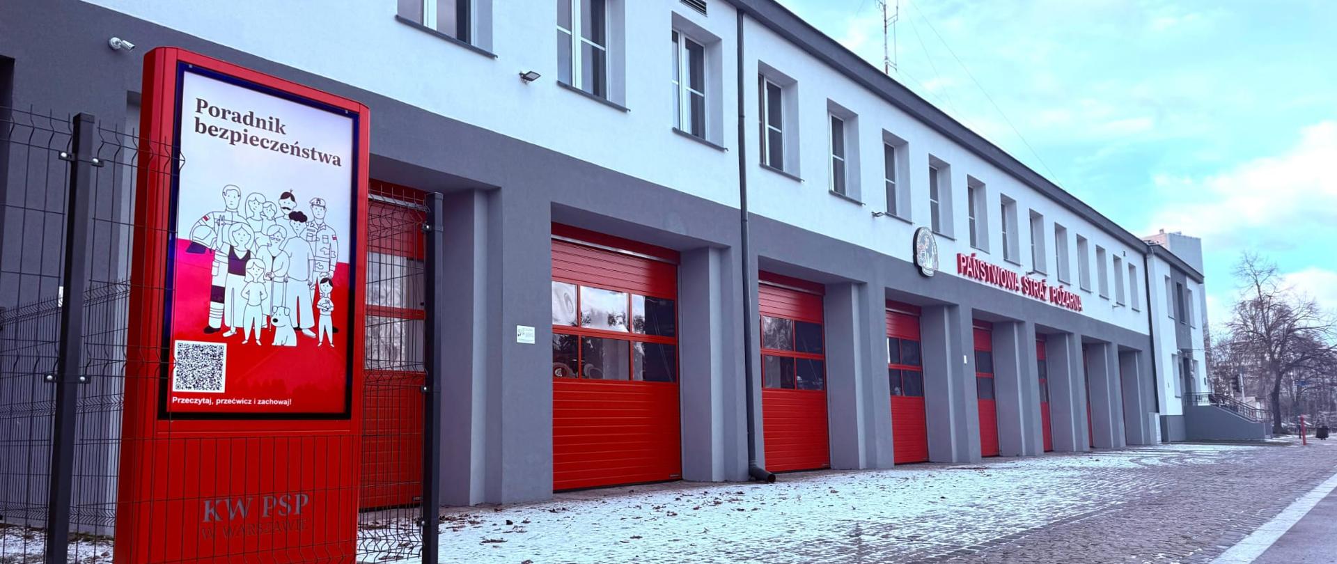 Monitor informacyjny w czerwonej obudowie stojący na tle budynku remizy strażackiej. 