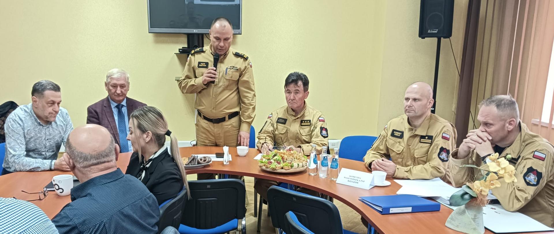 Zdjęcie zrobione w sali konferencyjnej, podczas ćwiczeń aplikacyjnych. Na sali siedzą uczestnicy ćwiczeń, wśród nich oficerowie Państwowej Straży Pożarnej. Jeden z uczestników przemawia do mikrofonu.